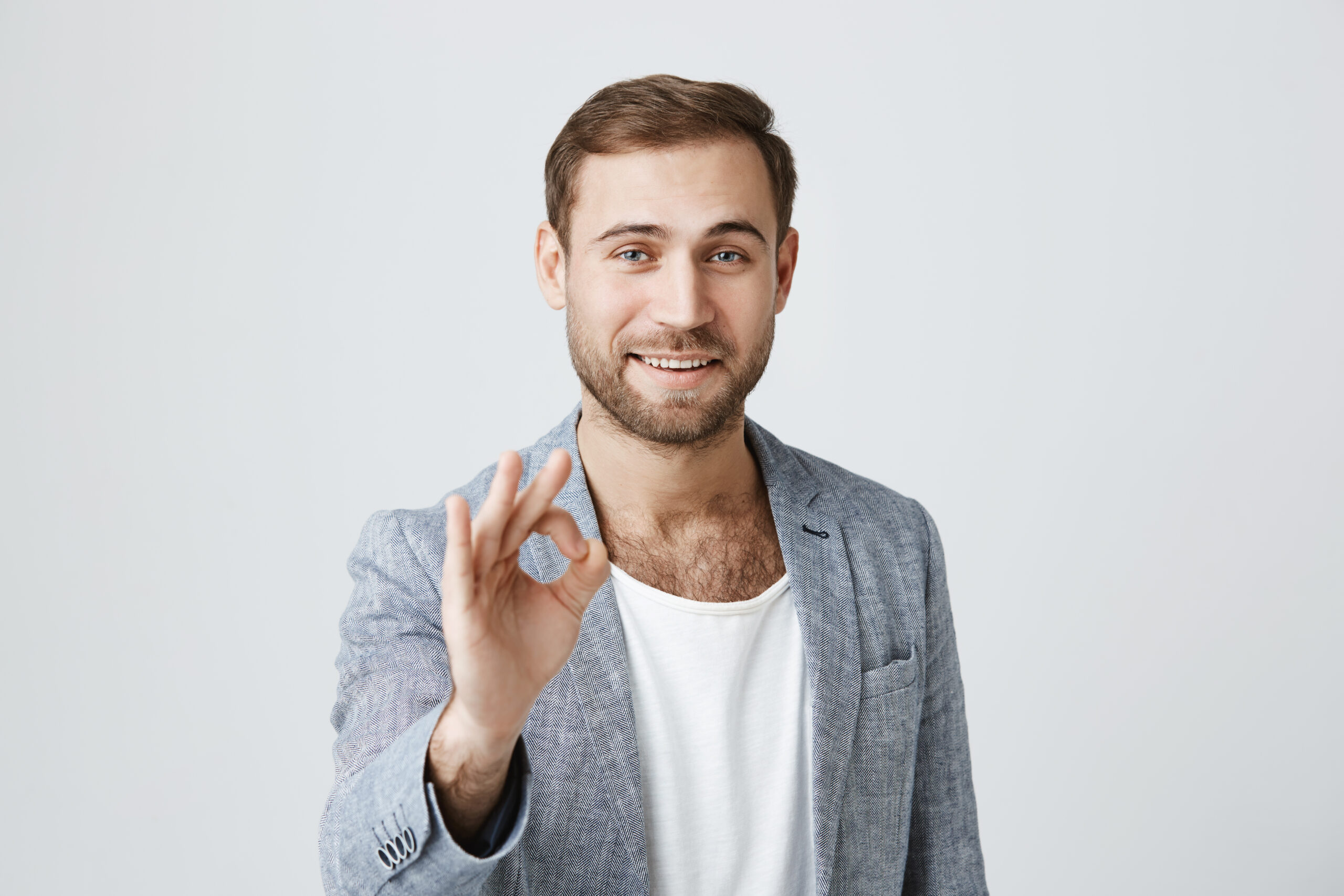 Positive bearded male wears trendy clothes makes ok gesture and smiles at camera, has happy expression, isolated against gray background. Optimistic male expresses his approval. Body language Amir A